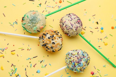 Photo of Delicious confectionery. Sweet cake pops and sprinkles on pale yellow background, flat lay