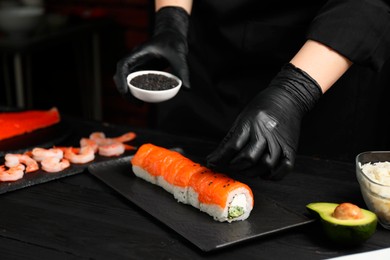 Photo of Chef in gloves adding sesame seeds onto tasty sushi rolls at black wooden table, closeup