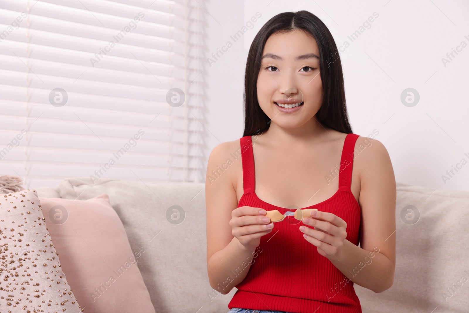 Photo of Asian woman holding tasty fortune cookie with prediction on sofa at home, space for text