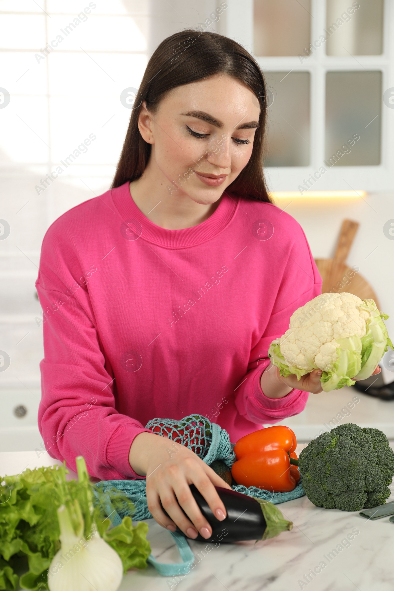 Photo of Woman taking vegetables out from string bag at light marble table in kitchen