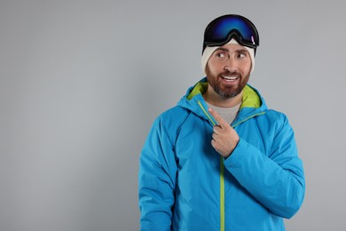 Photo of Winter sports. Happy man in ski suit and goggles pointing at something on gray background, space for text