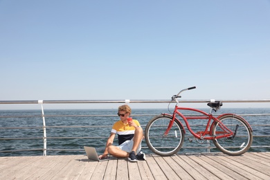 Attractive man with laptop and bike near sea on sunny day, space for text