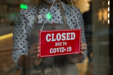 Photo of Woman in mask putting red sign with words Closed Due To Covid-19 hanging onto glass door, closeup