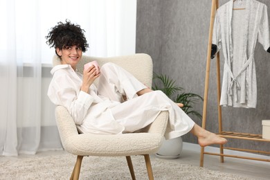 Photo of Beautiful young woman in stylish pyjama with cup of drink on armchair at home