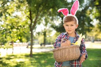 Cute little boy with bunny ears and basket of Easter eggs in park. Space for text