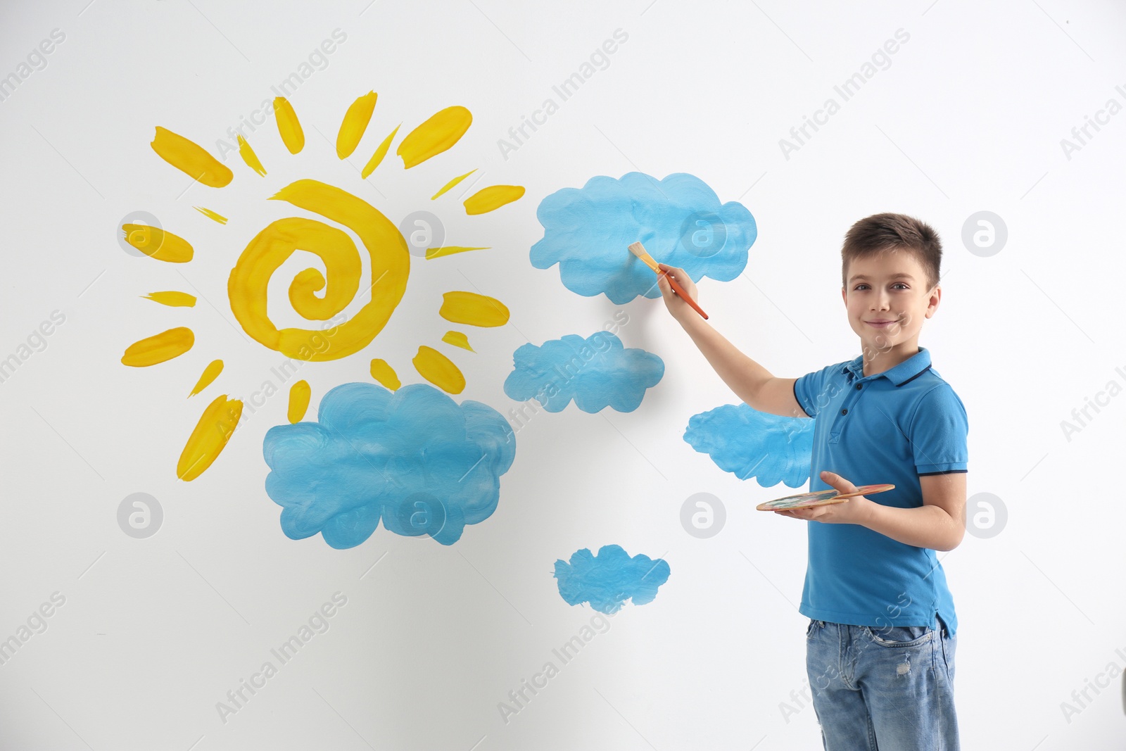 Image of Little boy drawing sun and clouds on white wall 