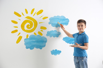 Image of Little boy drawing sun and clouds on white wall 