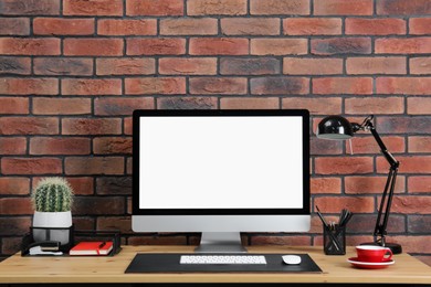 Stylish workplace with computer, lamp and stationery on wooden table near brick wall