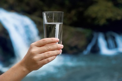 Photo of Woman holding glass of fresh water near waterfall outdoors, closeup. Space for text