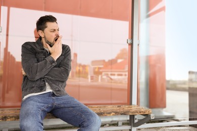 Sleepy man yawning at public transport stop outdoors. Space for text