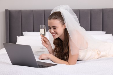 Photo of Happy bride with glass of sparkling wine and laptop on bed in bedroom
