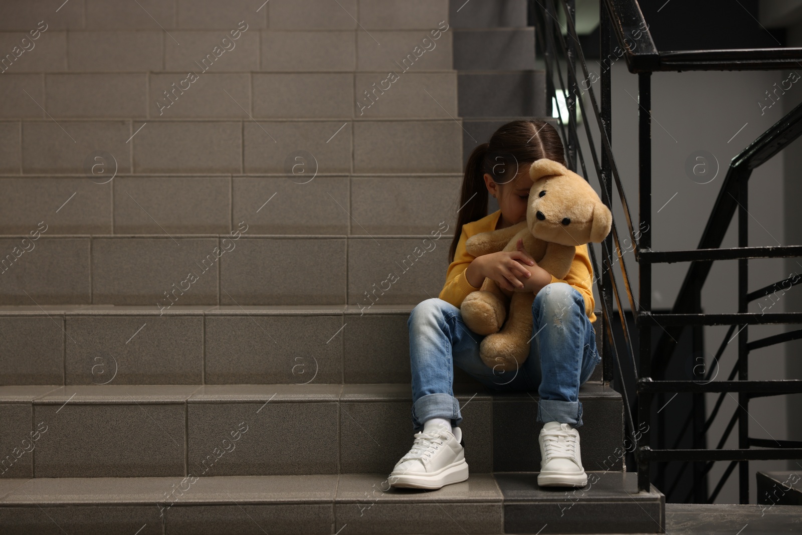 Photo of Child abuse. Upset little girl with teddy bear sitting on stairs indoors, space for text