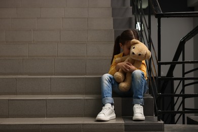 Photo of Child abuse. Upset little girl with teddy bear sitting on stairs indoors, space for text
