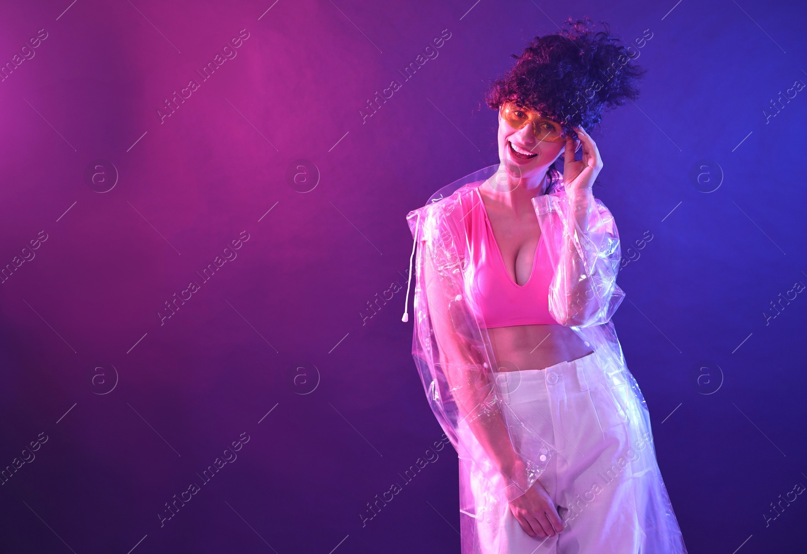 Photo of Beautiful young woman in transparent coat and sunglasses posing on color background in neon lights. Space for text