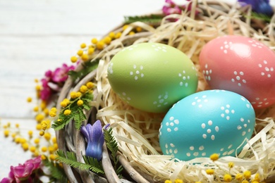 Photo of Wicker nest with painted Easter eggs and flowers on table, closeup