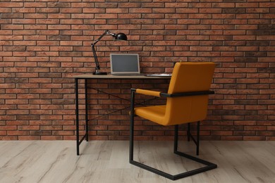 Comfortable office chair and desk with modern laptop near brick wall. Interior design