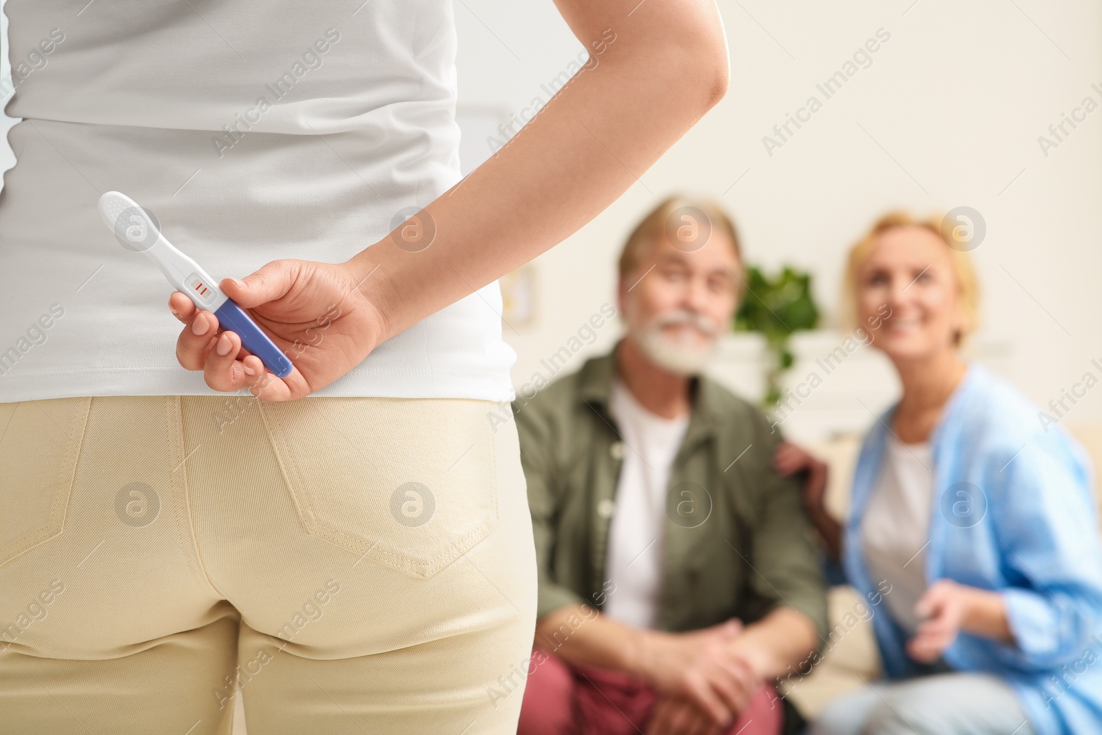 Photo of Woman holding pregnancy test behind her back near parents indoors, focus on hand. Grandparents' reaction to future grandson