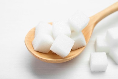 Photo of Many sugar cubes and wooden spoon on white table, closeup
