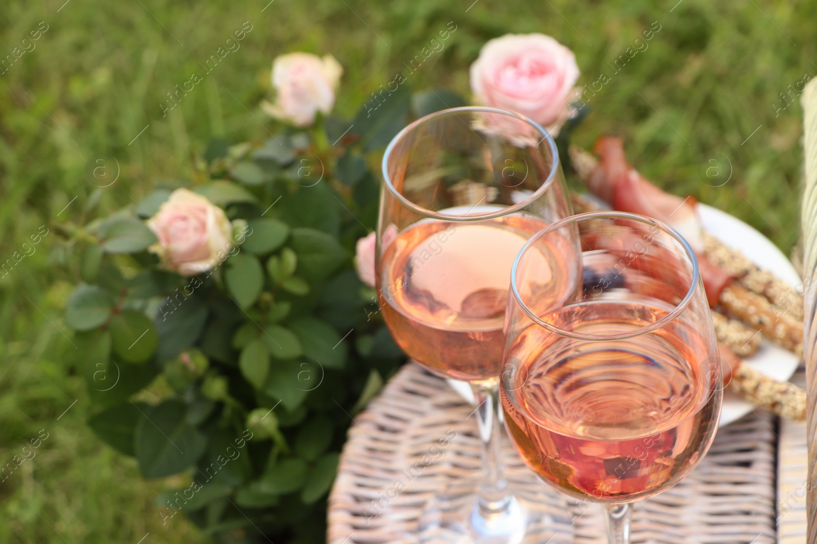 Photo of Flowers near glasses of delicious rose wine and food on picnic basket outdoors, closeup