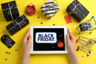 Photo of Woman with tablet surrounded by gifts on yellow background, top view. Black Friday sale