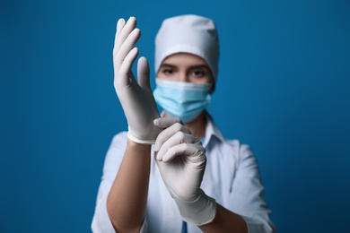 Doctor in protective mask putting on medical gloves against blue background