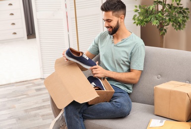 Young man opening parcel with shoes on sofa at home