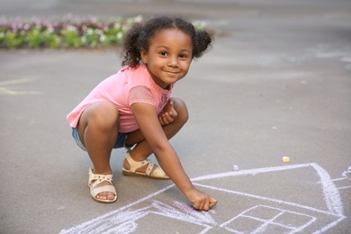 Little African-American child drawing house with chalk on asphalt