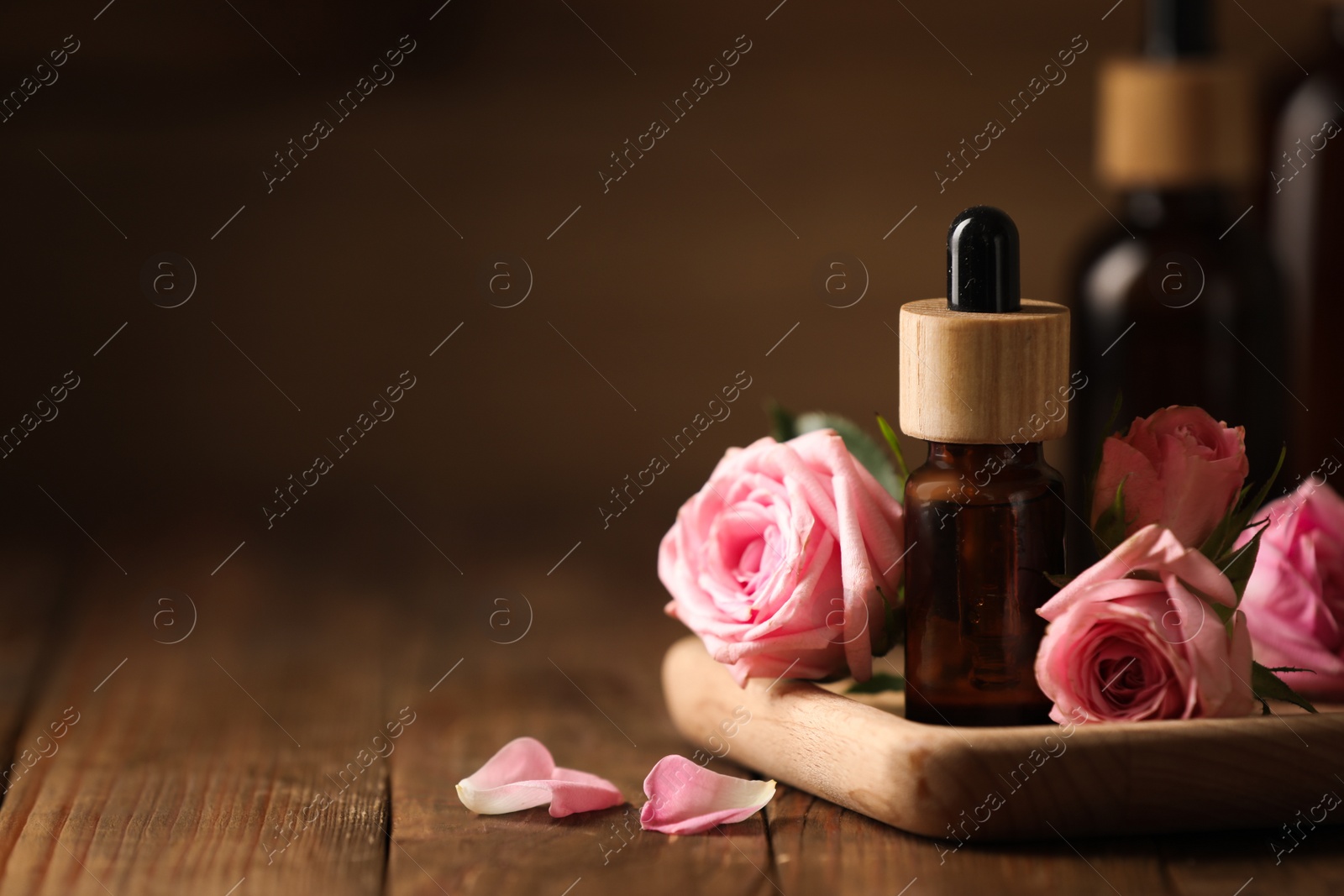 Photo of Bottles of essential rose oil and flowers on wooden table, space for text