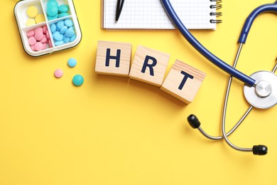 Image of Wooden cubes with abbreviation HRT, pills, stethoscope and notebook on yellow background, flat lay. Hormone Replacement Therapy