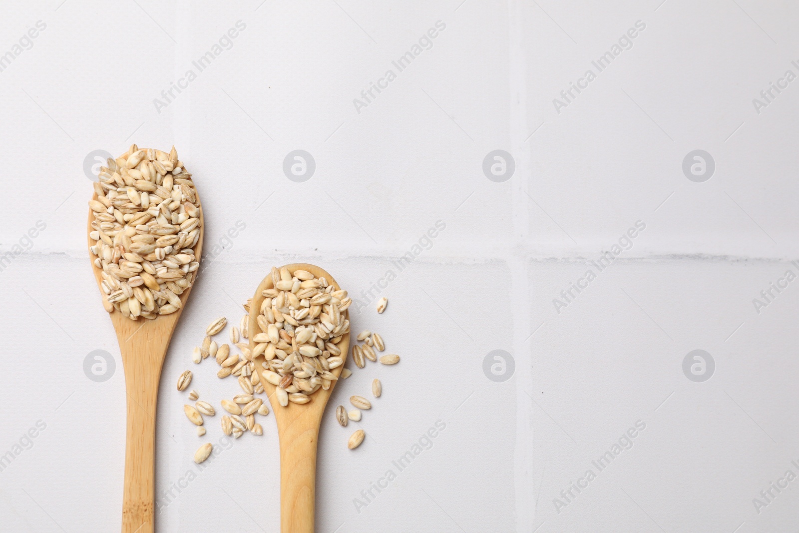 Photo of Wooden spoons with dry pearl barley on white tiled table, top view. Space for text