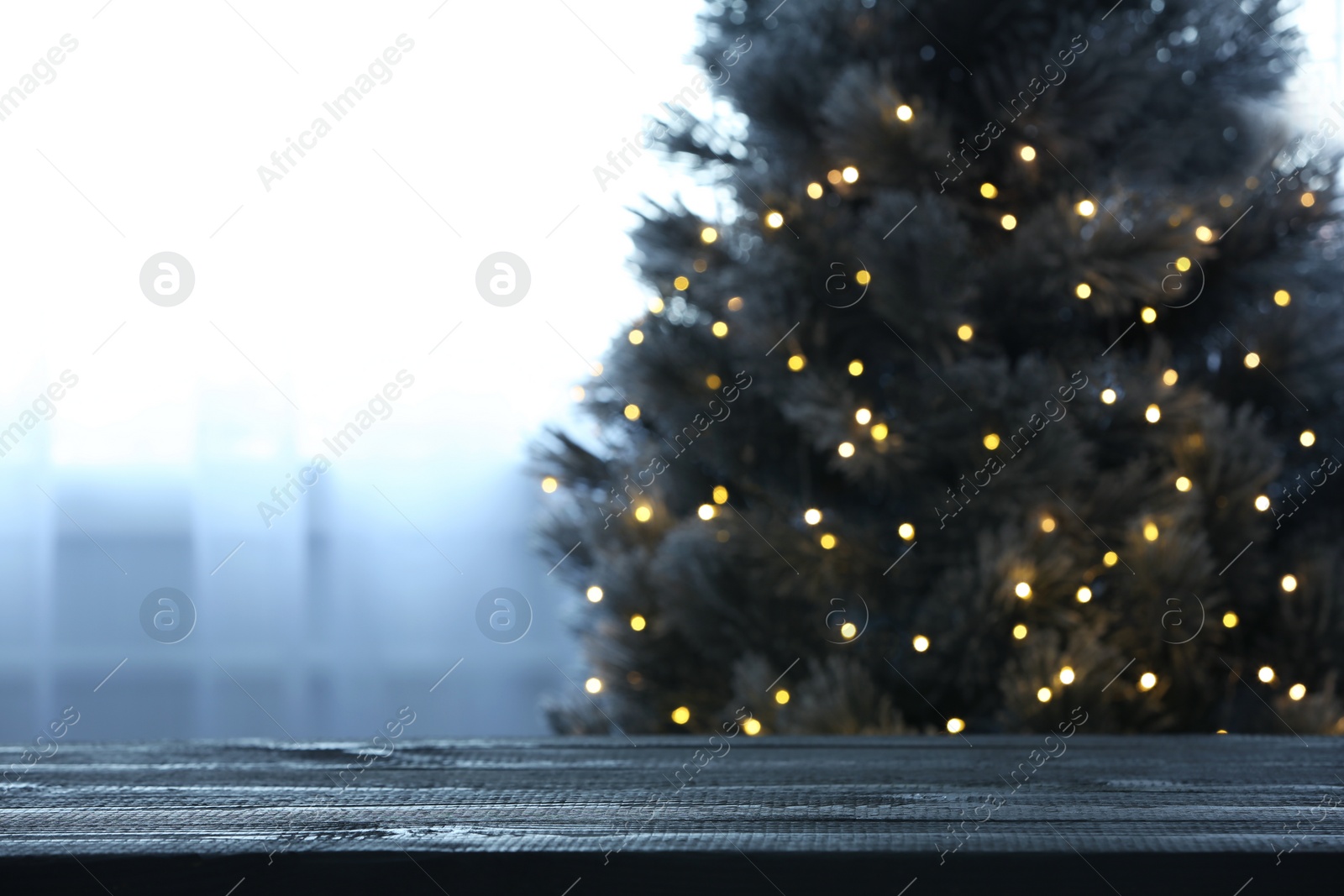 Photo of Blurred view of beautiful Christmas tree with yellow lights near window indoors, focus on wooden table. Space for text