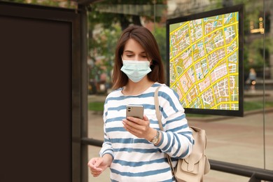 Image of Young woman in protective mask waiting for public transport at bus stop