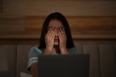 Photo of Young woman with nomophobia rubbing her eyes while using laptop at home late in evening. Insomnia concept