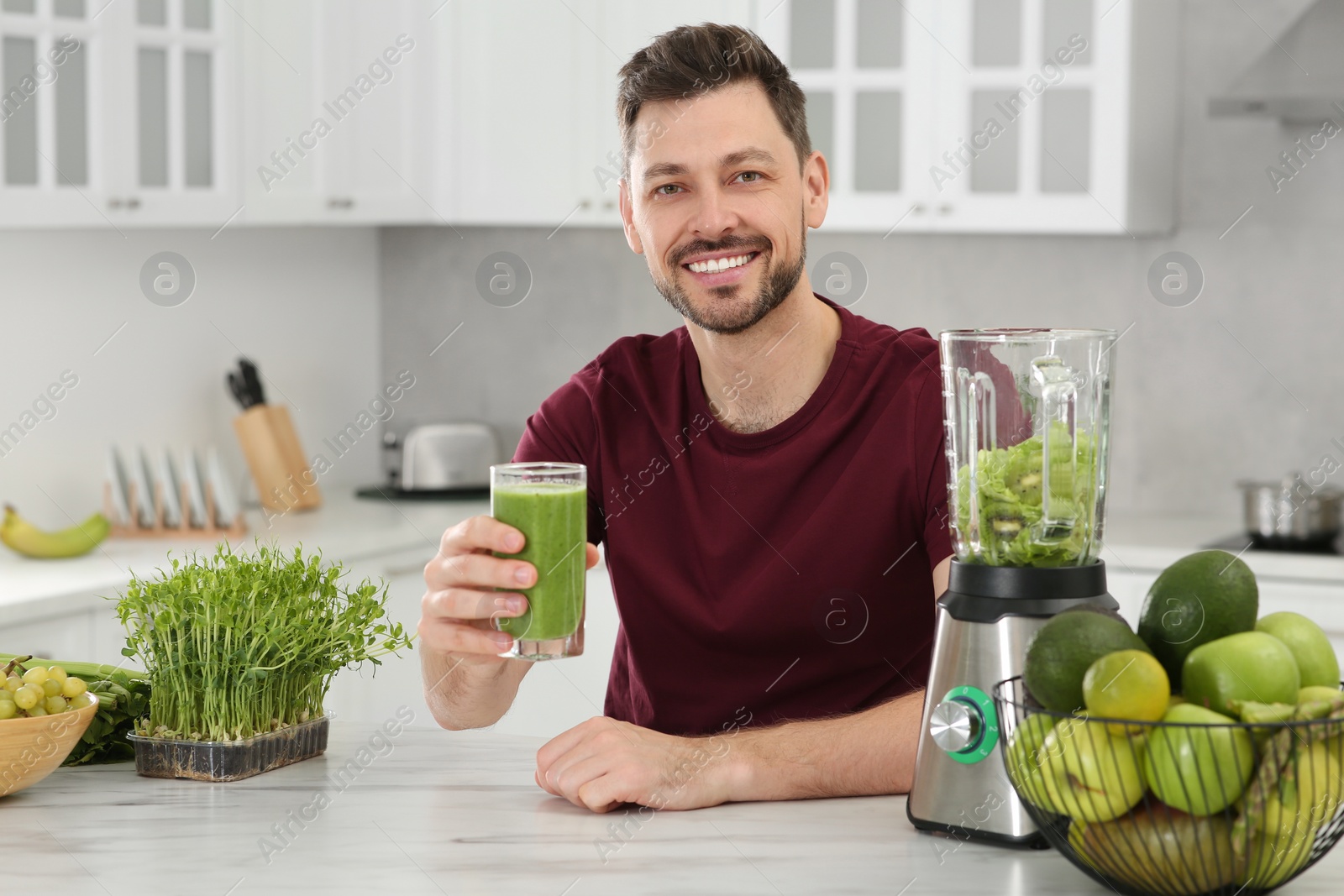 Photo of Happy man holding glass of delicious smoothie at white marble table in kitchen