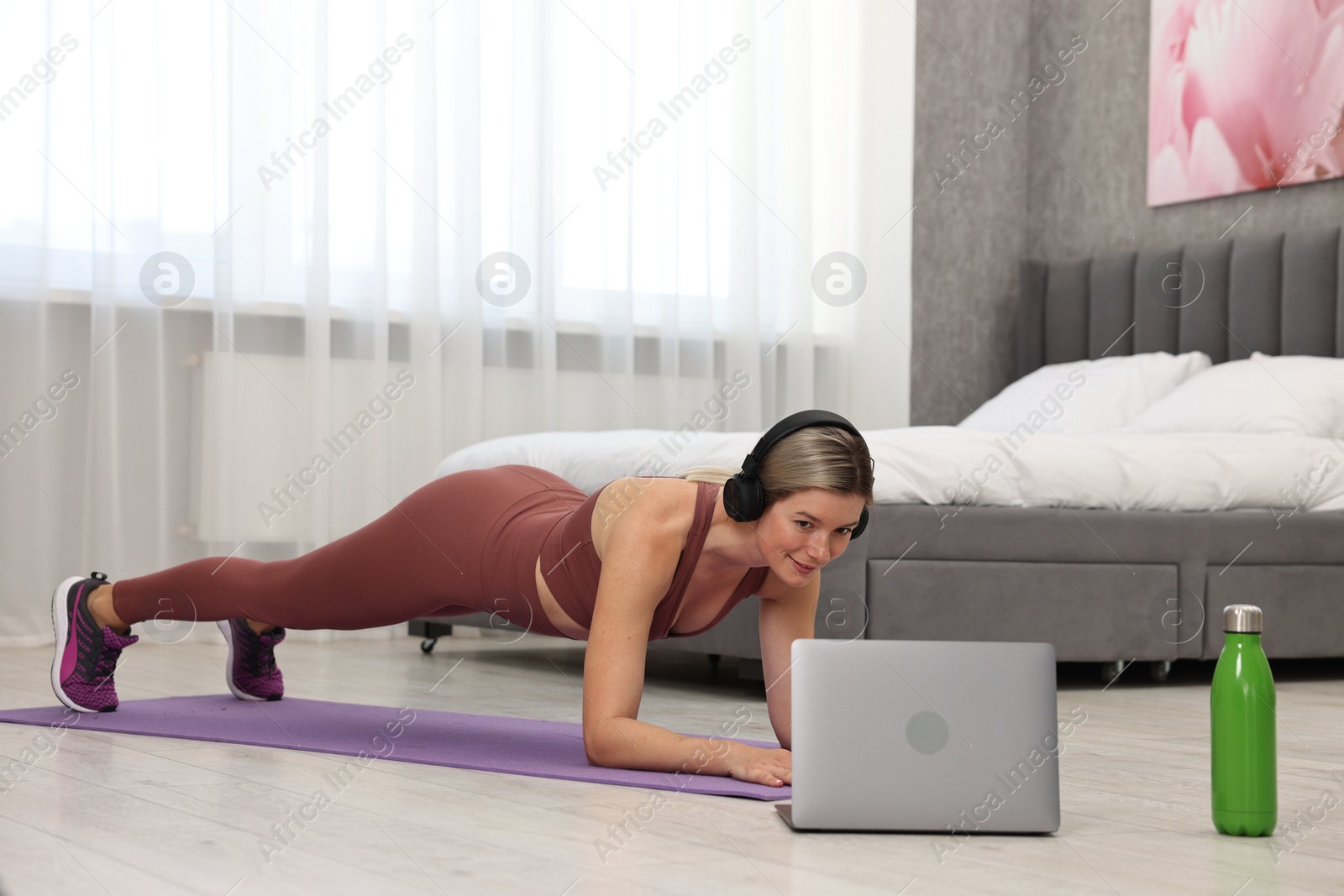 Photo of Online fitness trainer. Woman in headphones doing plank exercise near laptop at home