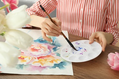 Photo of Woman painting flowers with watercolor at wooden table, closeup. Creative artwork
