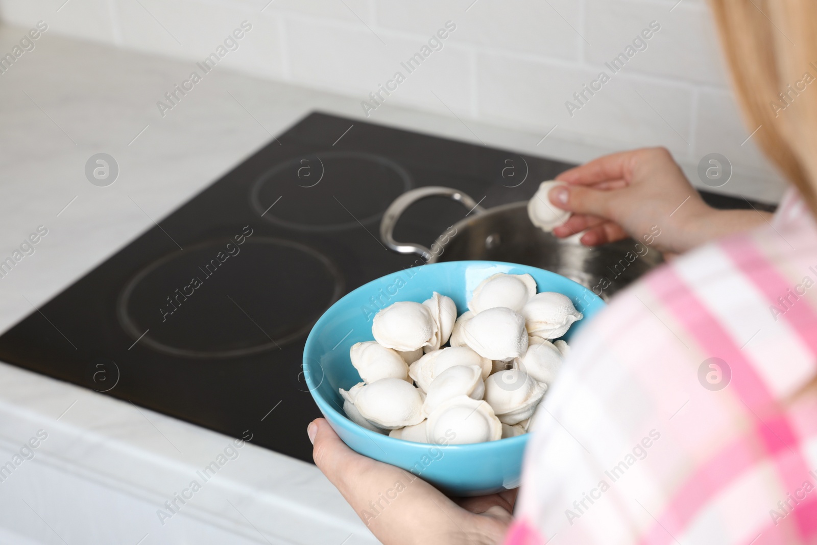 Photo of Woman putting frozen dumplings into saucepan with boiling water on cooktop in kitchen, closeup