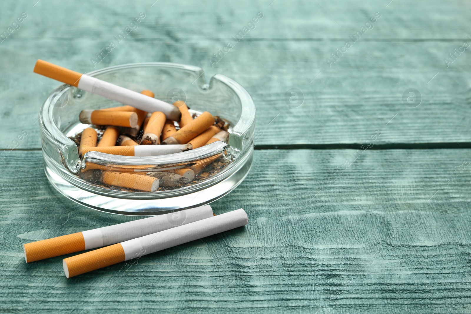 Photo of Glass ashtray with cigarette stubs on light blue wooden table. Space for text