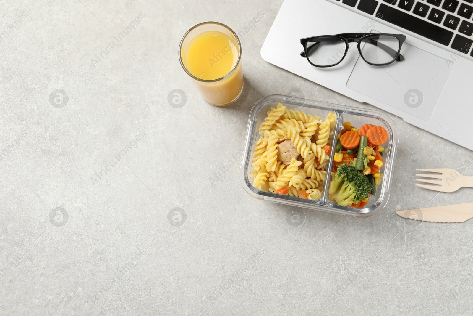 Photo of Container of tasty food, laptop, glasses, cutlery and glass of juice on light grey table, flat lay with space for text. Business lunch