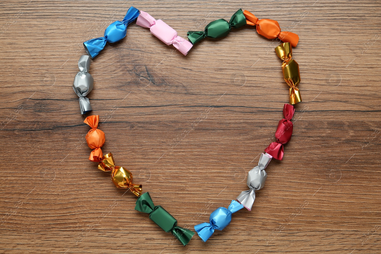 Photo of Heart shaped frame of candies in colorful wrappers on wooden table, flat lay. Space for text