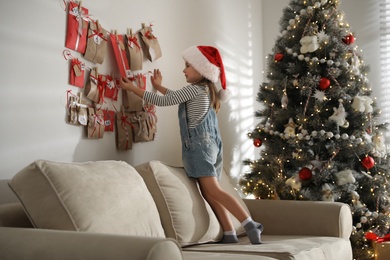 Cute little girl in Santa hat taking gift from Christmas advent calendar at home