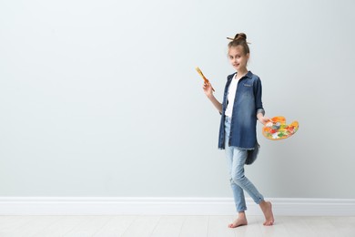 Little girl with brush and paint palette near light wall indoors. Space for text