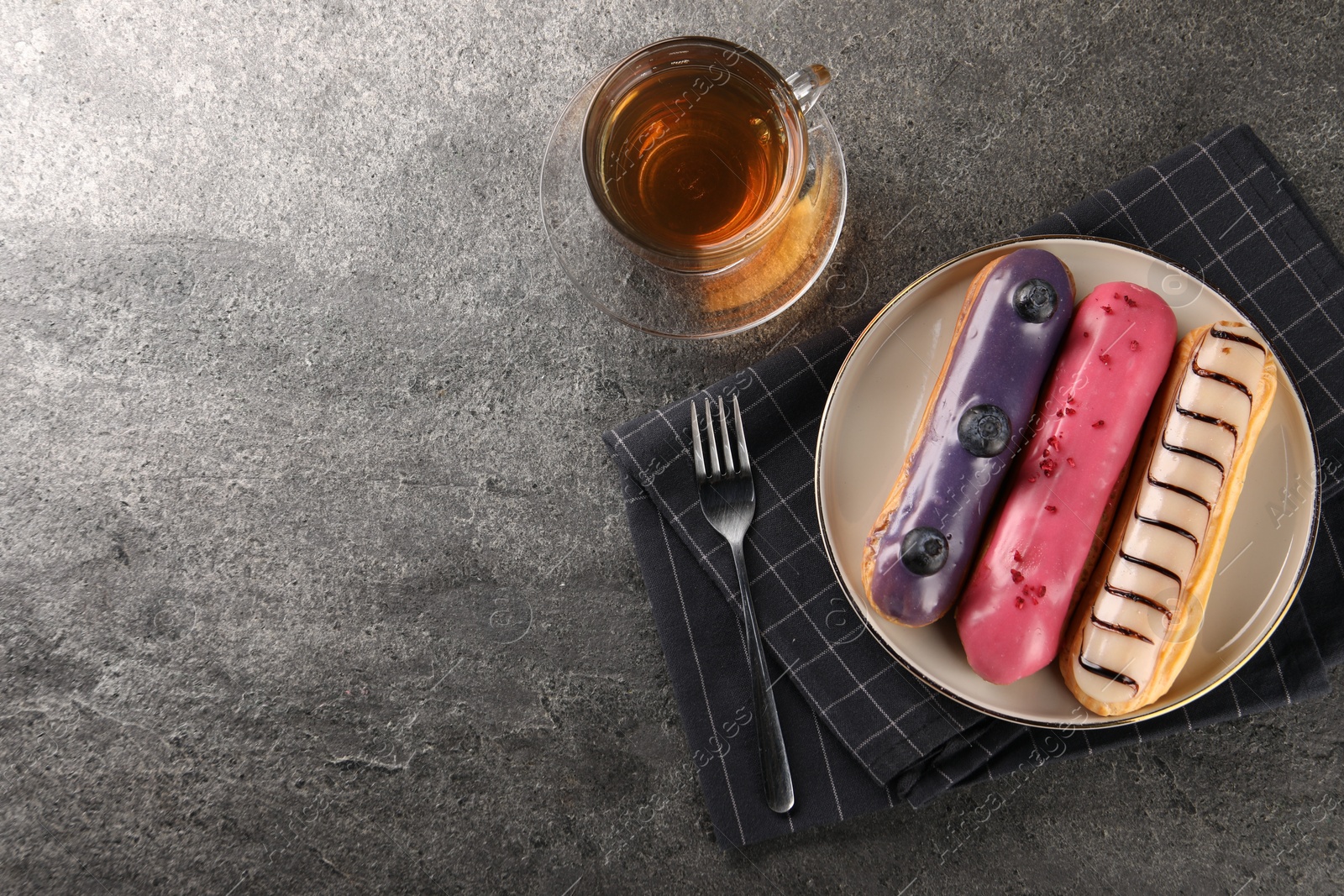 Photo of Different tasty glazed eclairs served with tea on grey textured table, flat lay. Space for text