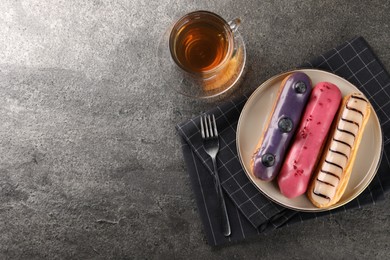 Different tasty glazed eclairs served with tea on grey textured table, flat lay. Space for text