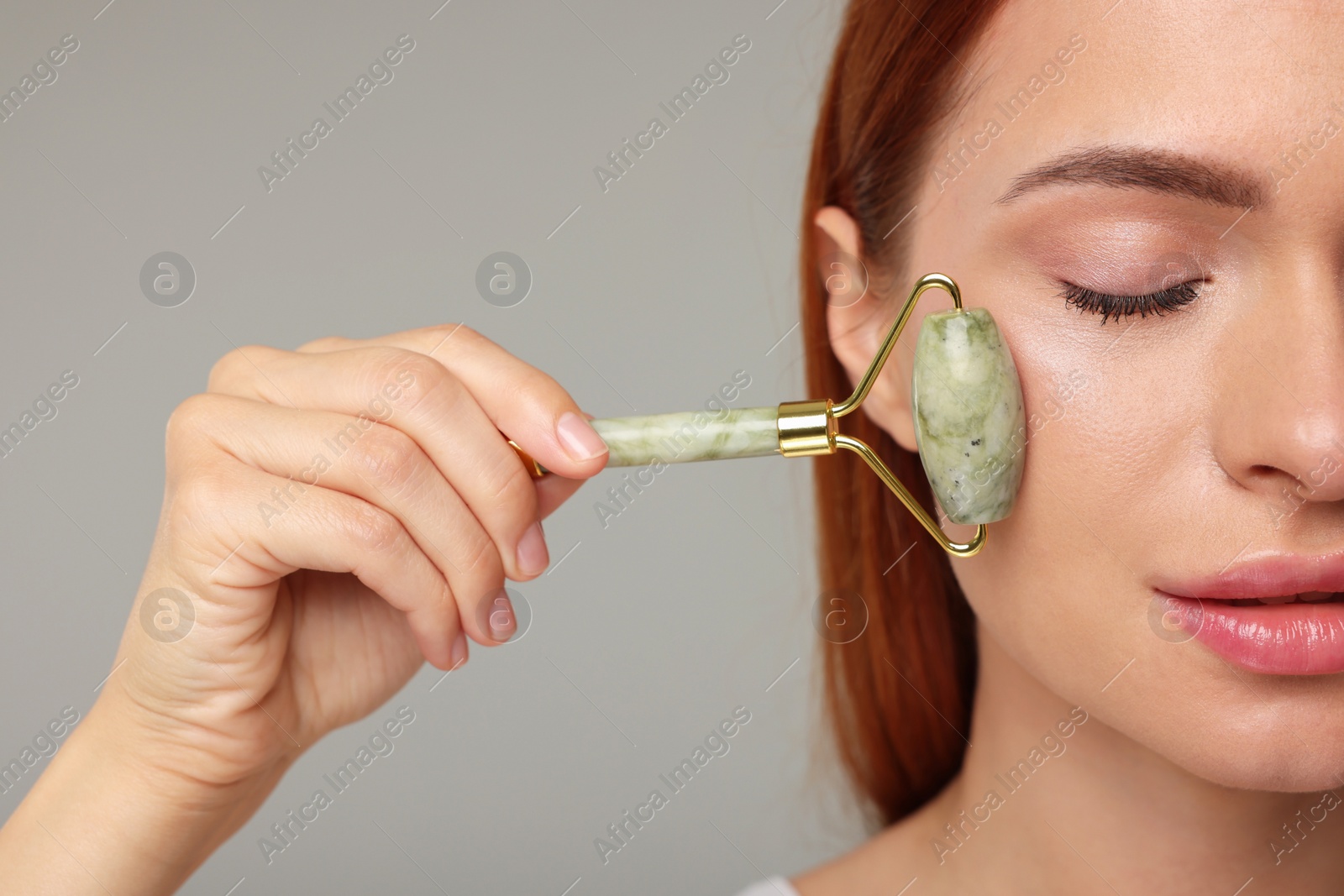 Photo of Young woman massaging her face with jade roller on grey background, closeup