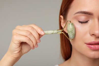 Photo of Young woman massaging her face with jade roller on grey background, closeup