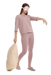 Young woman wearing pajamas, mask and slippers with pillow in sleepwalking state on white background