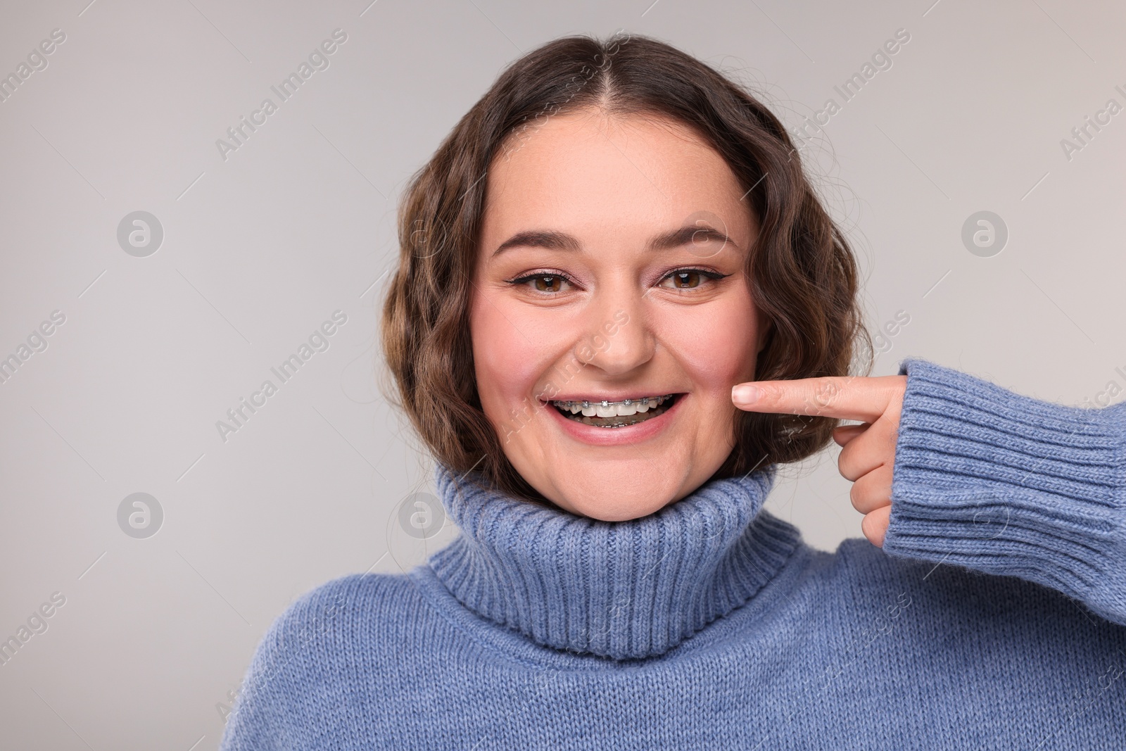 Photo of Happy woman in warm sweater pointing at braces on her teeth against grey background