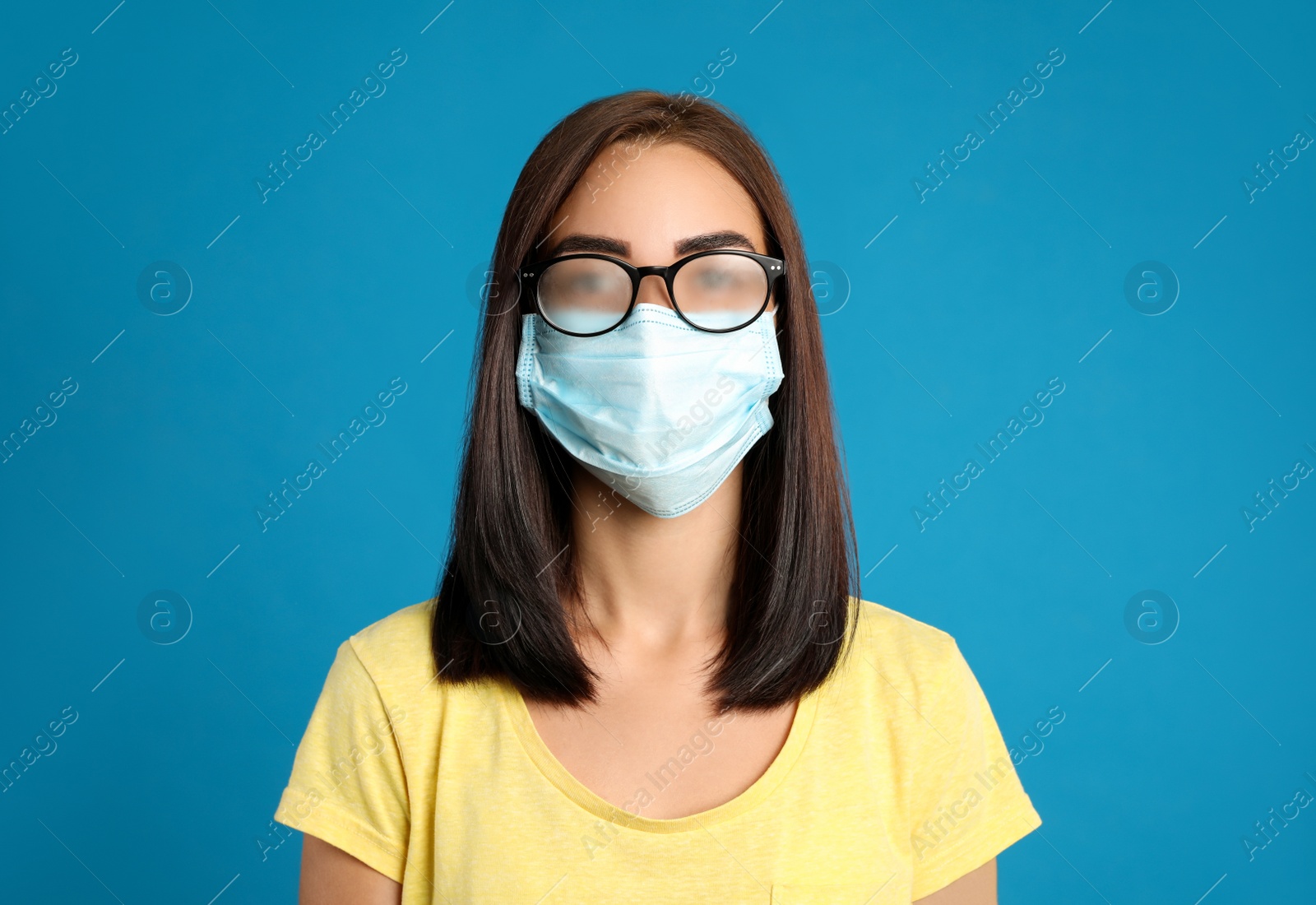 Photo of Young woman with foggy glasses caused by wearing disposable mask on blue background. Protective measure during coronavirus pandemic