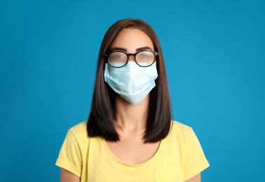 Photo of Young woman with foggy glasses caused by wearing disposable mask on blue background. Protective measure during coronavirus pandemic